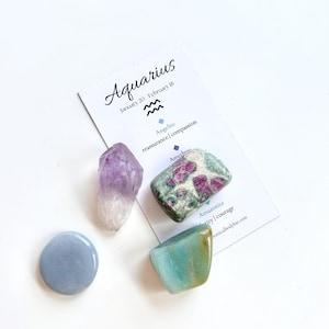 May include: An assortment of crystals and gemstones, including amethyst, amazonite, and angelite, arranged with a card that reads "Aquarius" and the dates January 20 - February 18. The gemstones are in shades of purple, blue, and green.