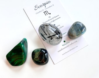 May include: A set of four tumbled stones, including black tourmaline, labradorite, malachite, and jasper. The stones are arranged on a white surface with a card that reads "Scorpio, October 23 - November 21, Tourmalinated Quartz, enlightenment, orite, wisdom, Jasper, vitali, Malachite, transformation | reflection, www.botanicalbodybar.com".