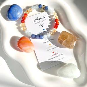 May include: A white ceramic dish with a blue, orange, and yellow beaded bracelet, a blue aventurine stone, an aquamarine stone, a carnelian stone, a citrine stone, and a white jade stone. The bracelet is labeled "Aries" with the astrological symbol for Aries. The stones are labeled with their names and properties.