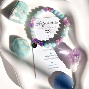 May include: Aquarius zodiac bracelet featuring a blend of light blue, pink, and purple beads. The bracelet is displayed on a white plate alongside several light blue and purple stones. The accompanying card reads "Aquarius, January 20 - February 18" with the astrological symbol for Aquarius. The card also details the stones used in the bracelet and their associated meanings.