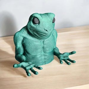 May include: A teal green 3D printed frog figurine with a human-like torso and arms. The frog has large, black eyes and a wide, toothy grin.