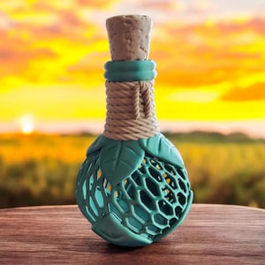 May include: A decorative bottle with a cork stopper. The bottle has a teal cage design with leaf accents and is wrapped with rope. The background is a blurred sunset over a field.