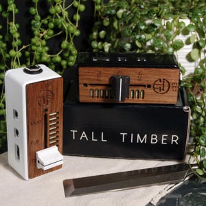 May include: Electronic devices with white and wood-grain panels, knobs, and sliders. The text "TALL TIMBER" is on a black box. A metal chisel is also present. The devices have a modern, industrial design.