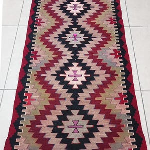 May include: A rectangular rug with a geometric pattern in shades of red, black, beige, and pink. The rug features a central diamond design with repeating patterns and a fringed edge. The rug's dimensions are not visible.