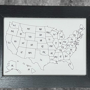 May include: A black framed map of the United States with each state labeled by its abbreviation. The map includes Alaska and Hawaii.