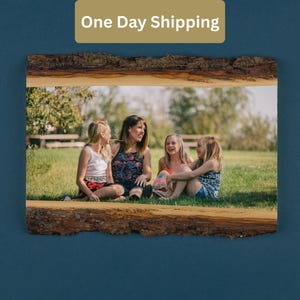 May include: A framed photograph with a natural wood bark border. The photo shows a group of four people sitting on the grass. The text "One Day Shipping" is in a gold rectangle at the top. The background is a grassy field with trees.