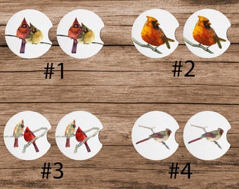 May include: Four different designs of round car coasters featuring watercolor paintings of cardinal birds. The first design shows two cardinals perched on a branch. The second design shows a single cardinal perched on a branch. The third design shows two cardinals perched on a branch. The fourth design shows a single cardinal perched on a branch.
