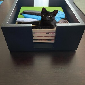 May include: A black kitten rests inside a black square coaster holder. The holder is on a dark brown wooden table. The kitten is lying on top of a stack of coasters, with a green and blue background.