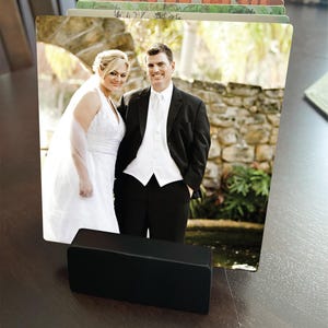 May include: A black photo holder displaying a wedding photo of a smiling couple. The bride wears a white wedding dress and veil, and the groom is in a black suit. The holder is rectangular and sits on a dark brown surface.
