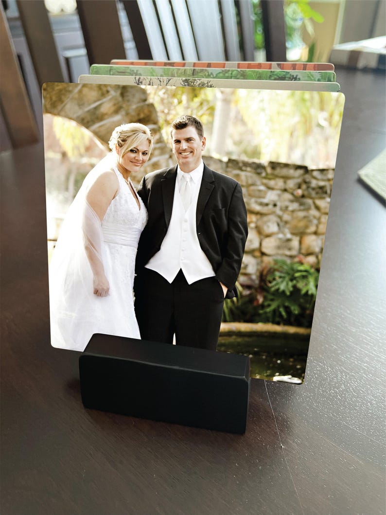 May include: A black photo holder displaying a wedding photo of a smiling couple. The bride wears a white wedding dress and veil, and the groom is in a black suit. The holder is rectangular and sits on a dark brown surface.