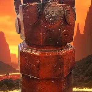 May include: A detailed, reddish-brown sculpture of a hand with a square design. The hand is set against a fiery, volcanic background, with a lava-like texture at the base. The sculpture has a textured, metallic appearance.