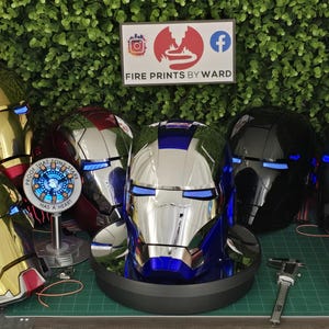 May include: A collection of Iron Man helmets in various metallic finishes including red and gold, blue and silver, and matte black, featuring glowing blue eye slits. Two circular arc reactor replicas are displayed alongside a metal caliper tool.