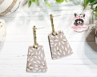 May include: A pair of dangle earrings with a light brown cork-like material with a white leaf pattern. The earrings have a gold-colored bamboo-like stem.