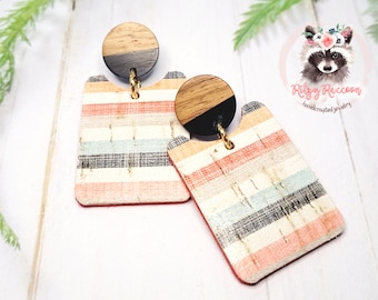 May include: A pair of striped cork earrings with a wood and black circle dangle. The earrings are handmade and feature a light pink, white, and blue striped pattern.