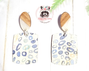 May include: A pair of dangle earrings with a white and brown oval top and a square bottom with a blue and white abstract pattern. The earrings are handmade and feature a silver chain.
