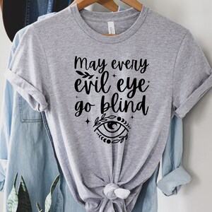 May include: A light gray t-shirt with the text "May every evil eye go blind" in black script. The design includes an eye symbol with floral accents. The shirt is hanging on a wooden hanger with a blue denim shirt underneath.