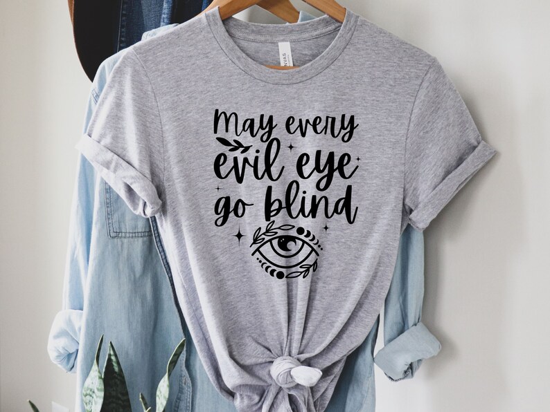 May include: A light gray t-shirt with the text "May every evil eye go blind" in black script. The design includes an eye symbol with floral accents. The shirt is hanging on a wooden hanger with a blue denim shirt underneath.