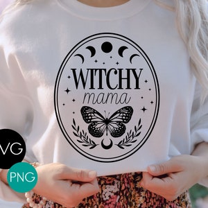 K&ouml;nnte beinhalten: Wei&szlig;es Sweatshirt mit einem schwarzen Grafikdesign. Das Design zeigt die Worte "Witchy Mama" in einem ovalen Rahmen, einen Schmetterling, Halbmonde und Sterne. Das Bild enth&auml;lt auch die Texte "SVG" und "PNG" in einem schwarzen und t&uuml;rkisfarbenen Kreis.