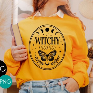 K&ouml;nnte beinhalten: Senfgelbes Sweatshirt mit schwarzem Aufdruck &bdquo;Witchy Mama&ldquo; in einem ovalen Rahmen. Der Rahmen enth&auml;lt Halbmonde, Sterne, einen Schmetterling und Bl&auml;tter. Das Bild enth&auml;lt auch SVG- und PNG-Dateityp-Symbole.
