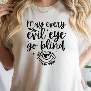 May include: A beige t-shirt with the phrase "May every evil eye go blind" in black script. The design includes an eye symbol with leaves and stars. The shirt is a casual wear item.