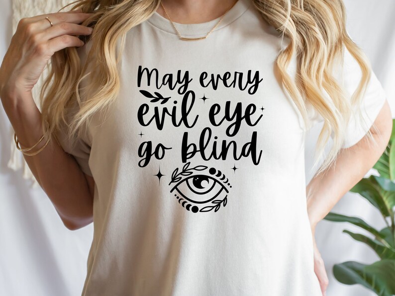 May include: A beige t-shirt with the phrase "May every evil eye go blind" in black script. The design includes an eye symbol with leaves and stars. The shirt is a casual wear item.