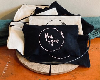 May include: A stack of black and white canvas pouches with zippers. The front pouch has the text "You do you" in a pink circle with the text "So you can be you" written around the circle.