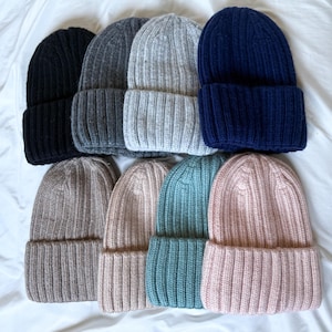 May include: A collection of eight ribbed knit beanies arranged in two rows. The top row features black, dark grey, light grey, and navy blue beanies. The bottom row includes tan, beige, teal, and light pink beanies, all with a folded cuff design.