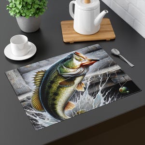 May include: A rectangular placemat featuring a detailed illustration of a largemouth bass leaping out of water. The fish is depicted with green, yellow, and black scales, with water splashing around it. A fishing lure hangs nearby. The background shows weathered wood.