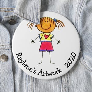May include: A white button with a cartoon drawing of a girl in a yellow shirt with a red heart and red shorts. The button has the text "Raylene's Artwork 2020" in a circular pattern around the edge. The button is pinned to a denim jacket.