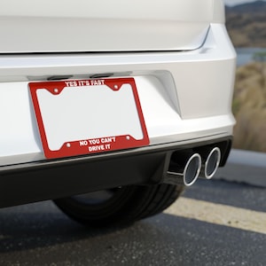Könnte beinhalten: Ein roter Kennzeichenrahmen mit dem Text "YES IT'S FAST" oben und "NO YOU CAN'T DRIVE IT" unten. Der Rahmen ist an der Rückseite eines weißen Autos in der Nähe der Auspuffrohre angebracht.