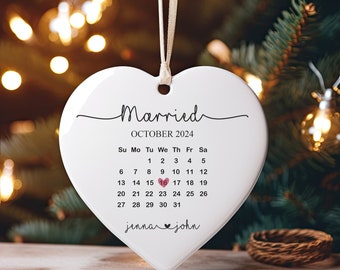 May include: White heart-shaped ceramic ornament with the words "Married" and "October 2024" printed on it. A calendar with the date "16" highlighted in pink is printed below the text. The names "jenna" and "john" are printed at the bottom of the ornament.