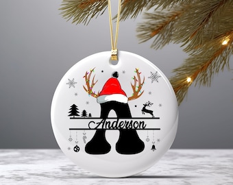 May include: White ceramic Christmas bauble featuring a black silhouette of a reindeer wearing a Santa hat. The reindeer's antlers are adorned with lights. The bauble also includes a black silhouette of a Christmas tree, a deer, and the text "Anderson".