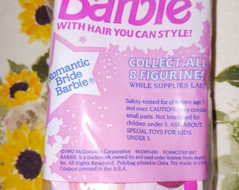 May include: A pink and white plastic bag with a Barbie doll inside. The bag has the words "Barbie with hair you can style!" and "Collect all 8 figurines while supplies last." The bag also has a copyright notice for 1992 McDonald's Corporation.