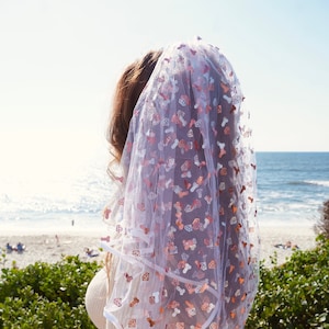 May include: A white bridal veil with a pink and white pattern of diamonds and other shapes. The veil is made of a sheer fabric and has a scalloped edge.