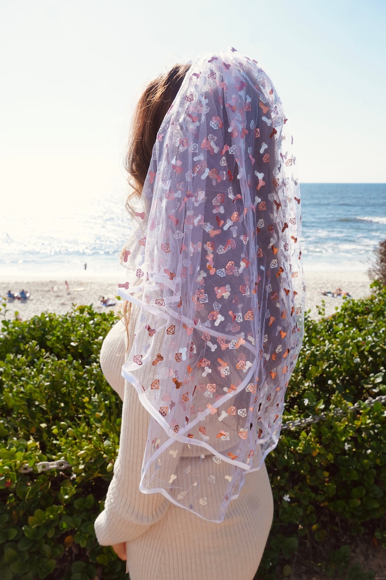 May include: A white bridal veil with a pink and white pattern of diamonds and other shapes. The veil is made of a sheer fabric and has a scalloped edge.