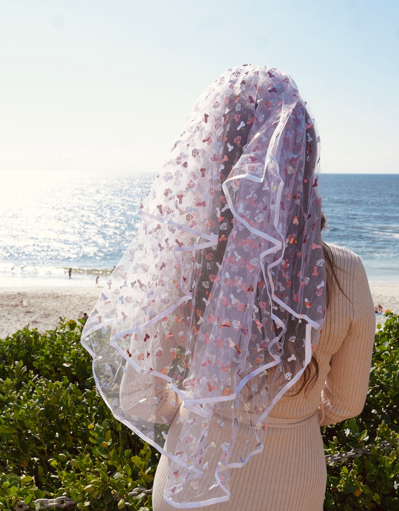 May include: A white bridal veil with a white satin trim and pink and white glitter hearts and other shapes.