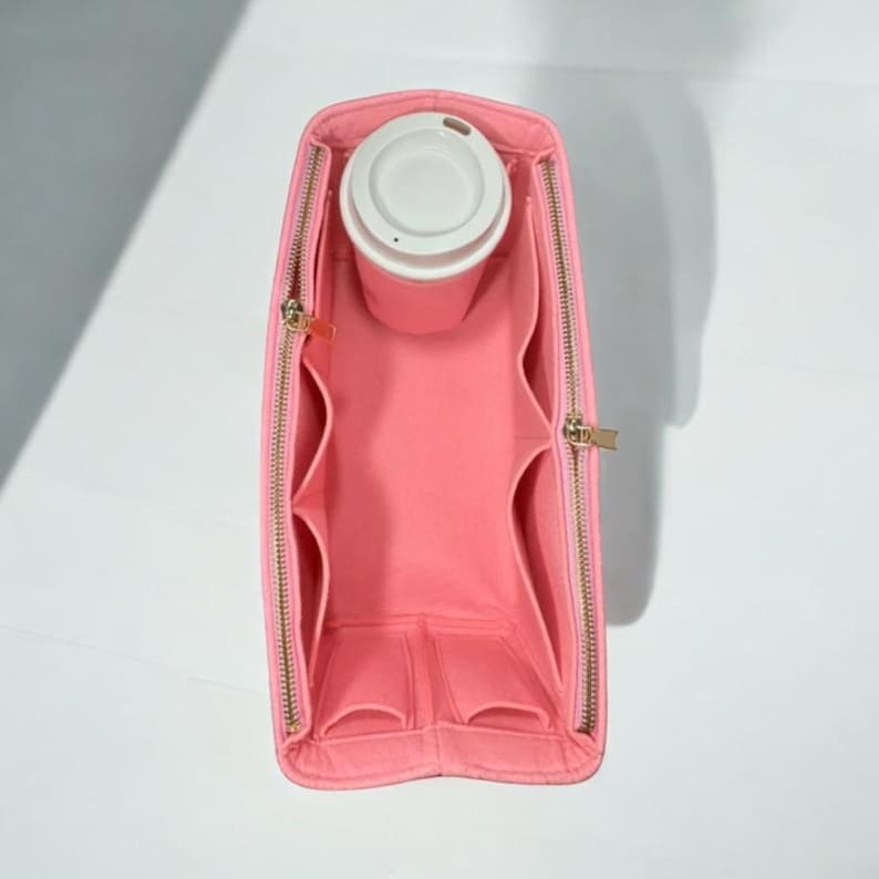 Puede incluir: Organizador de bolso de fieltro rosa con m&uacute;ltiples compartimentos y cierre de cremallera. El organizador tiene un espacio dedicado para una taza o una botella de agua.