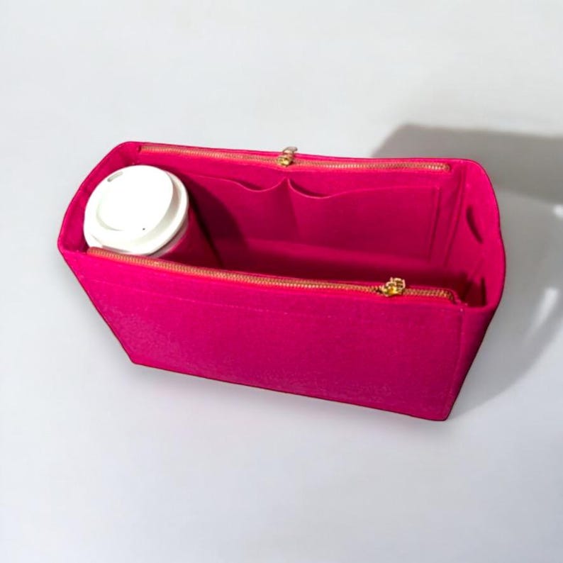 Puede incluir: Un organizador de bolso de fieltro rosa brillante con una cremallera dorada. El organizador tiene varios compartimentos y un bolsillo para una taza.