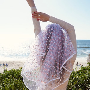 May include: A white bridal veil with a pink and white pattern of hearts and other shapes. The veil is made of tulle and has a white satin trim.