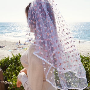 May include: A white tulle veil with pink and silver glitter hearts and other shapes. The veil is worn by a person standing on a beach.