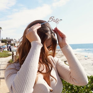 May include: A woman wearing a beige knitted sweater with buttons and a bride headband. She is standing in front of a beach and ocean.