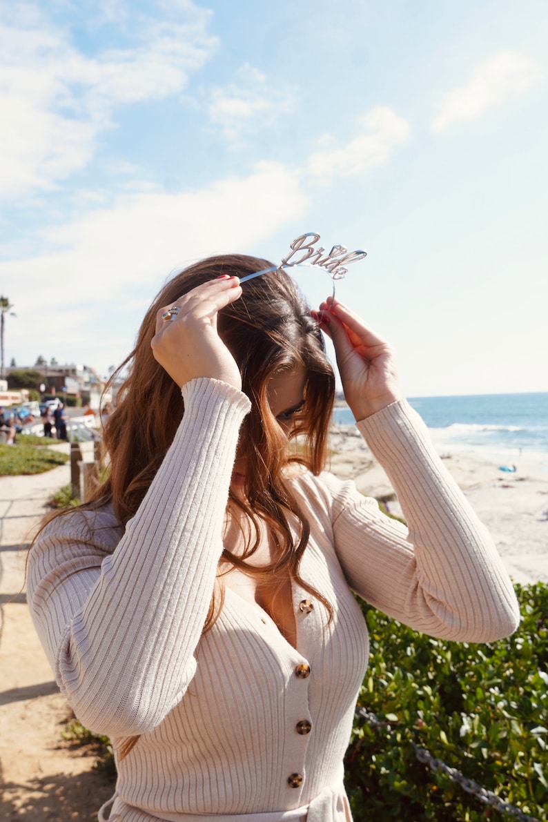 May include: A woman wearing a beige knitted sweater with buttons and a bride headband. She is standing in front of a beach and ocean.