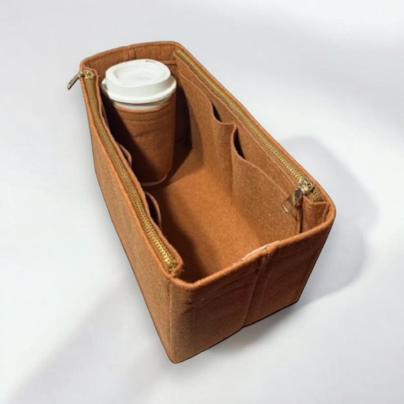 Puede incluir: Un organizador de bolso de fieltro marr&oacute;n con m&uacute;ltiples compartimentos y cierre de cremallera. El organizador est&aacute; dise&ntilde;ado para caber dentro de un bolso m&aacute;s grande e incluye un bolsillo dedicado para una taza de caf&eacute;.
