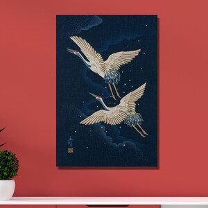 Op de afbeelding: Een canvas print met twee kraanvogels in vlucht tegen een donkerblauwe achtergrond met wolken. De kraanvogels hebben witte en gouden vleugels met blauwe accenten. Het kunstwerk wordt op een rode muur getoond.