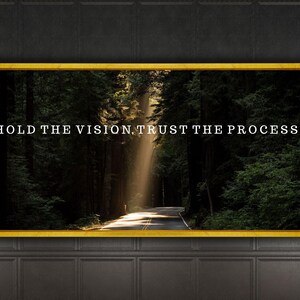 Puede incluir: Una cita motivacional, "Hold the vision, trust the process", se muestra en un lienzo enmarcado con un marco dorado. La imagen de fondo es un camino forestal con un rayo de sol que brilla a través de los árboles.