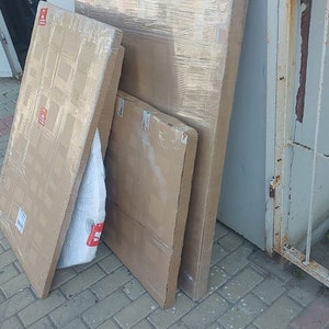 May include: Three cardboard boxes stacked on top of each other. The boxes are wrapped in clear plastic and have a brown and white pattern. The boxes are sitting on a gray brick surface.