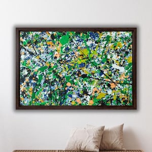 Puede incluir: Pintura abstracta en un marco marrón oscuro. La obra presenta una vibrante mezcla de colores, incluyendo verde, azul, naranja, amarillo, negro y blanco, aplicados en un estilo salpicado. La pintura se exhibe sobre dos cojines beige.