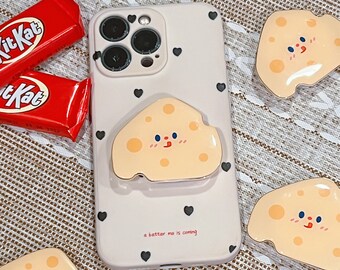 May include: A white phone case with a black heart pattern and a cartoon cheese phone grip. The phone case has a camera lens with three black circles. The phone grip is a cartoon cheese with a smiling face and a pink blush. The text "a better me is coming" is printed on the phone case.