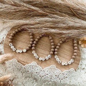 Peut inclure: Trois bracelets en perles avec les mots "Mum of" et un charme en forme de cœur. Les bracelets sont beiges avec des lettres blanches et des accents dorés.