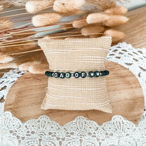 Peut inclure: Un bracelet en perles noires avec des lettres blanches qui épellent "DAD OF" et un petit charme en forme de cœur. Le bracelet est présenté sur un petit coussin beige.